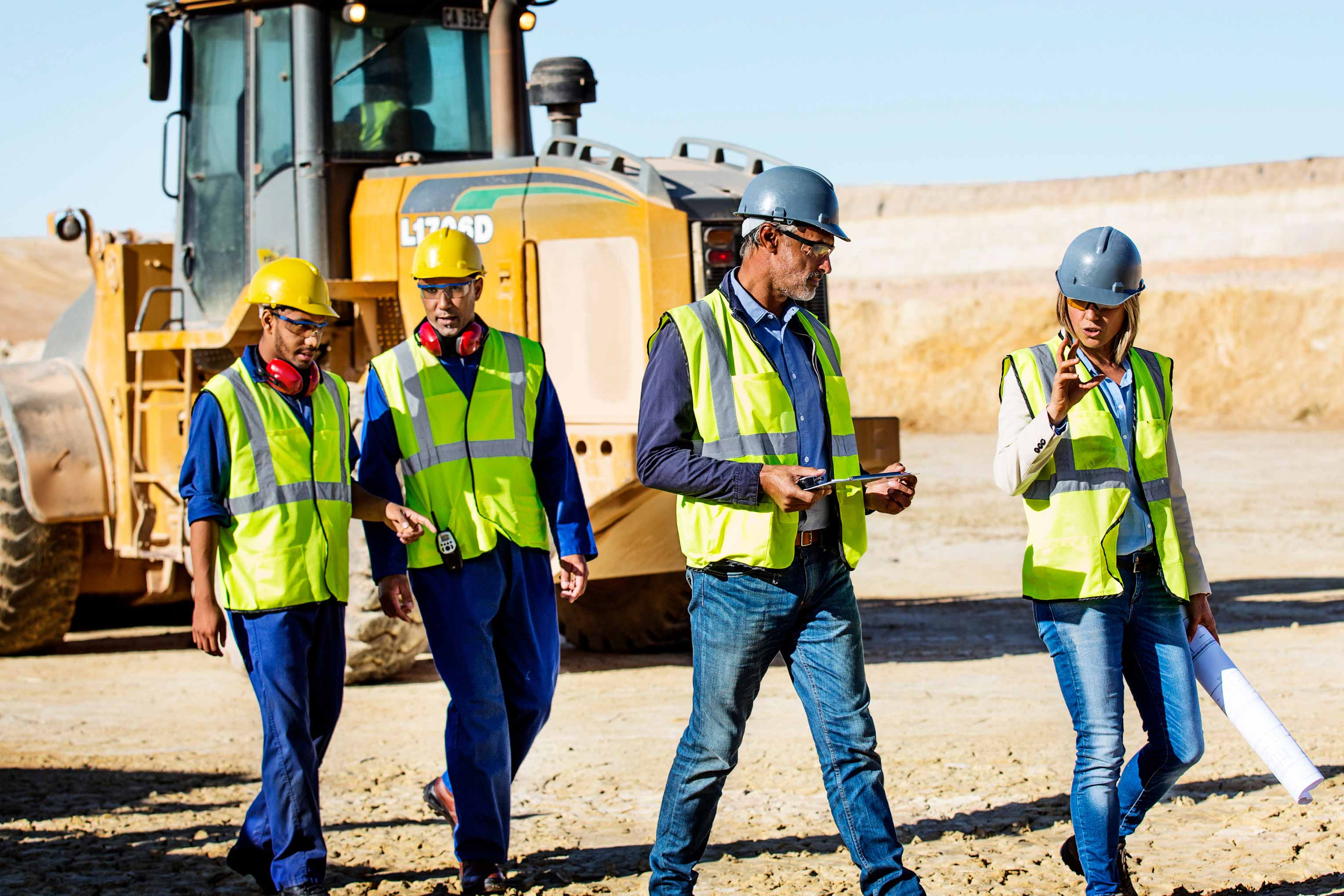 A construction team tours a job site for a future data center project. They were safety vests, hard hats and dark work clothes as they walk in front of large construction equipment. 