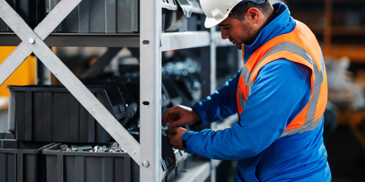 A male worker in a warehouse is examining goods on a shelf. He is wearing a high-visibility vest and a hard hat, ensuring safety while performing his duties. The background shows rows of shelves filled with various items, indicating a busy and organized warehouse environment, highlighting effective storeroom management.