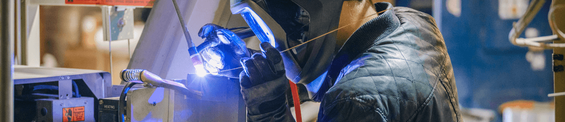 A welder wearing dark and heavy protective clothing, light brown gloves and welding helmet, welds a seam on a metal frame.