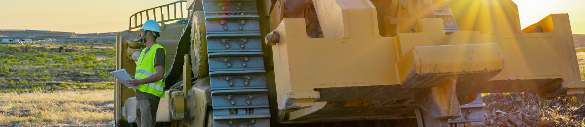 On a flat stretch of land during a sunrise, a man wearing a white hard hat, yellow safety vest and grey pants and shoes faces away from a parked bulldozer.