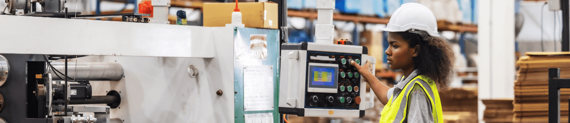 In a manufacturing plant, a woman wearing a white hardhat, orange safety vest, grey work shirt and dark blue pants. She stands at the end of an industrial stitching machine and is pressing a button on the interface. Search the latest machine operator jobs with Aerotek.
