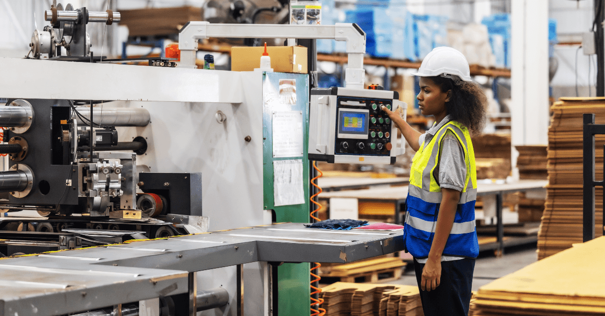 In a manufacturing plant, a woman wearing a white hardhat, orange safety vest, grey work shirt and dark blue pants. She stands at the end of an industrial stitching machine and is pressing a button on the interface. Find the latest machine operator jobs with Aerotek.