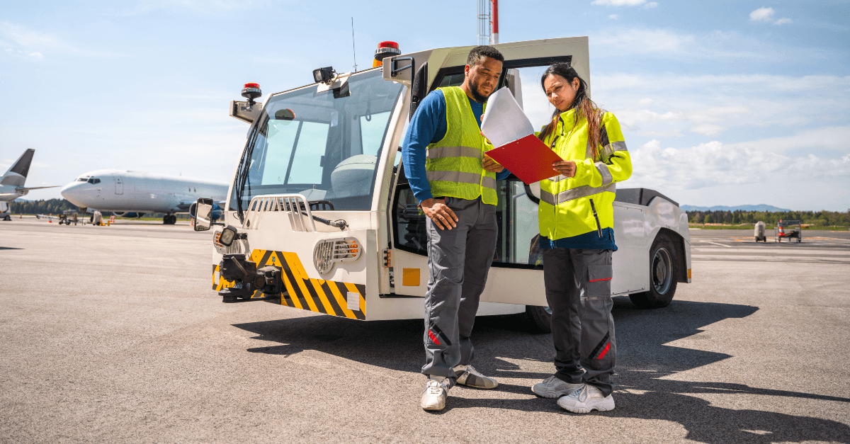 On an airport runway, two workers stand in front of a small truck and review information on a clipboard. The man on the left wears a yellow safety vest, long-sleeve blue shirt, grey pants and white shoes. The woman on the right wears a yellow safety jacket, grey pants and white shoes. 