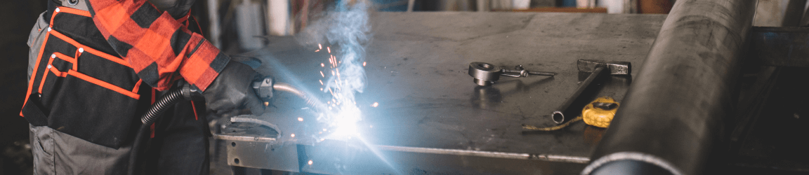 In a factory, a welder wearing heavy duty face protection and heavy protective clothing welds while looking down at their workspace.