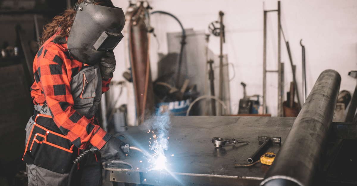 In a factory, a welder wearing heavy duty face protection and heavy protective clothing welds while looking down at their workspace. 