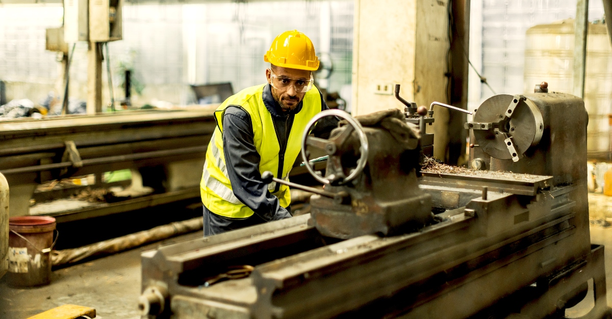 A skilled millwright in a yellow hard hat and reflective vest is operating a large industrial lathe machine. Metal shavings are visible, indicating active metalworking. The workshop setting is dimly lit with various manufacturing equipment in the background.