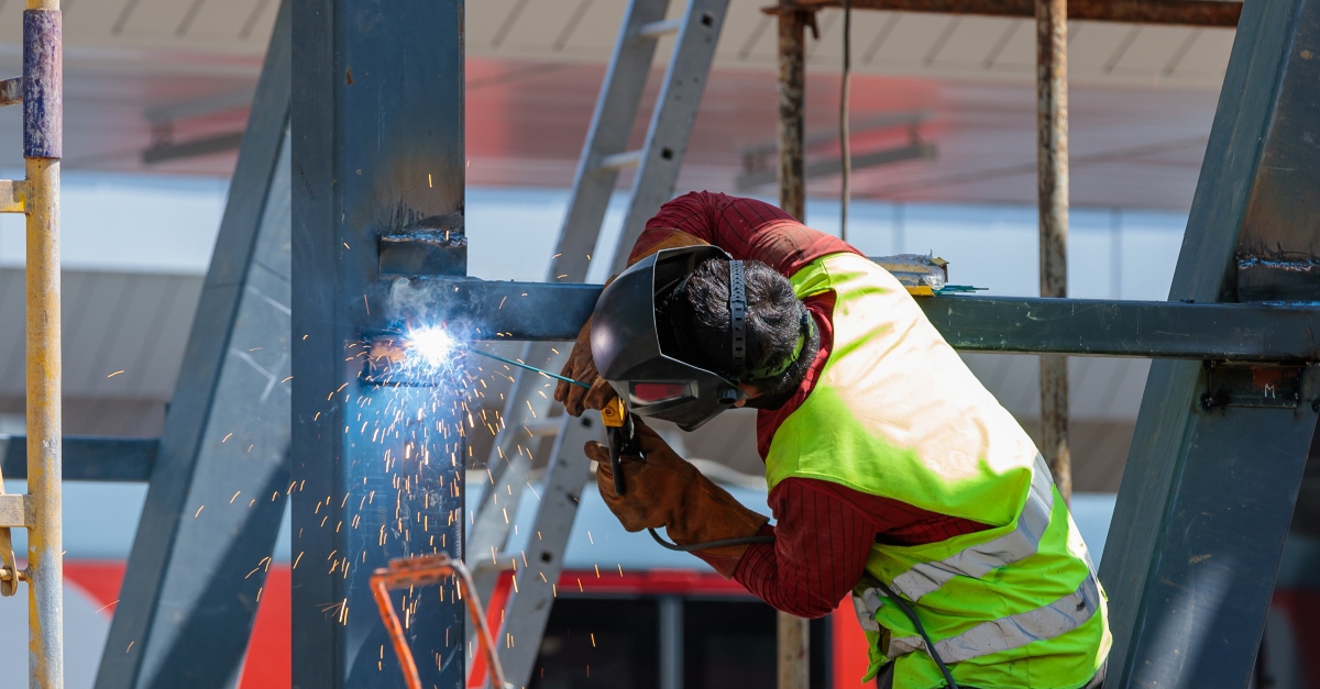 A man welder fastens iron girders with a welding machine at a construction site. Sparks fly as he works, wearing protective gear including a helmet and gloves. The background shows the framework of a building under construction.