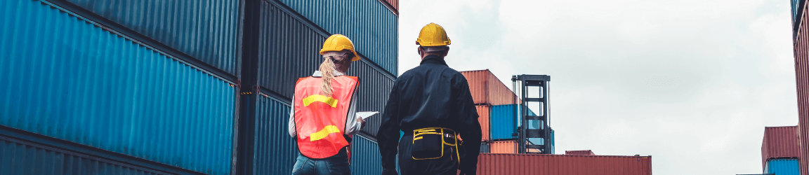 With their backs facing the camera, a man and woman wearing hard hats walk through a shipping container yard.