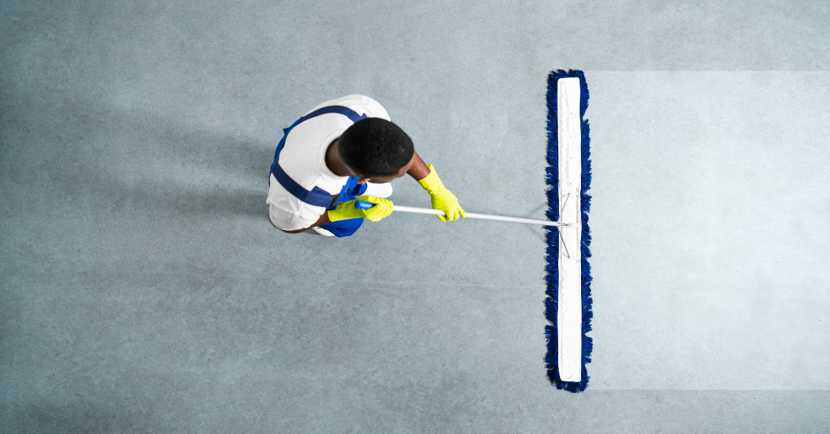 A top-down view shows a man wearing a white shirt, yellow gloves and blue overalls as he uses a large dust broom on a grey floor. The broom leaves a path of clean floor as he pulls it towards himself.