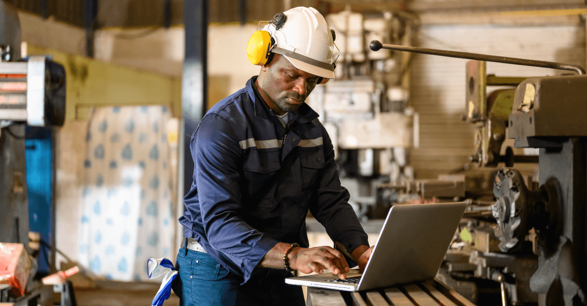 A male technician in a blue shirt, wearing a hardhat and protective earmuffs, works in a shop