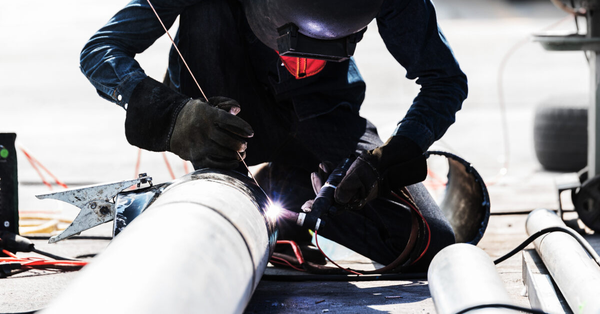 A pipefitter perches over a metal pipe while welding the end closest to them. The welder wears a long-sleeve dark blue shirt and dark grey gloves. They are also wearing a tan and grey welding hood. 