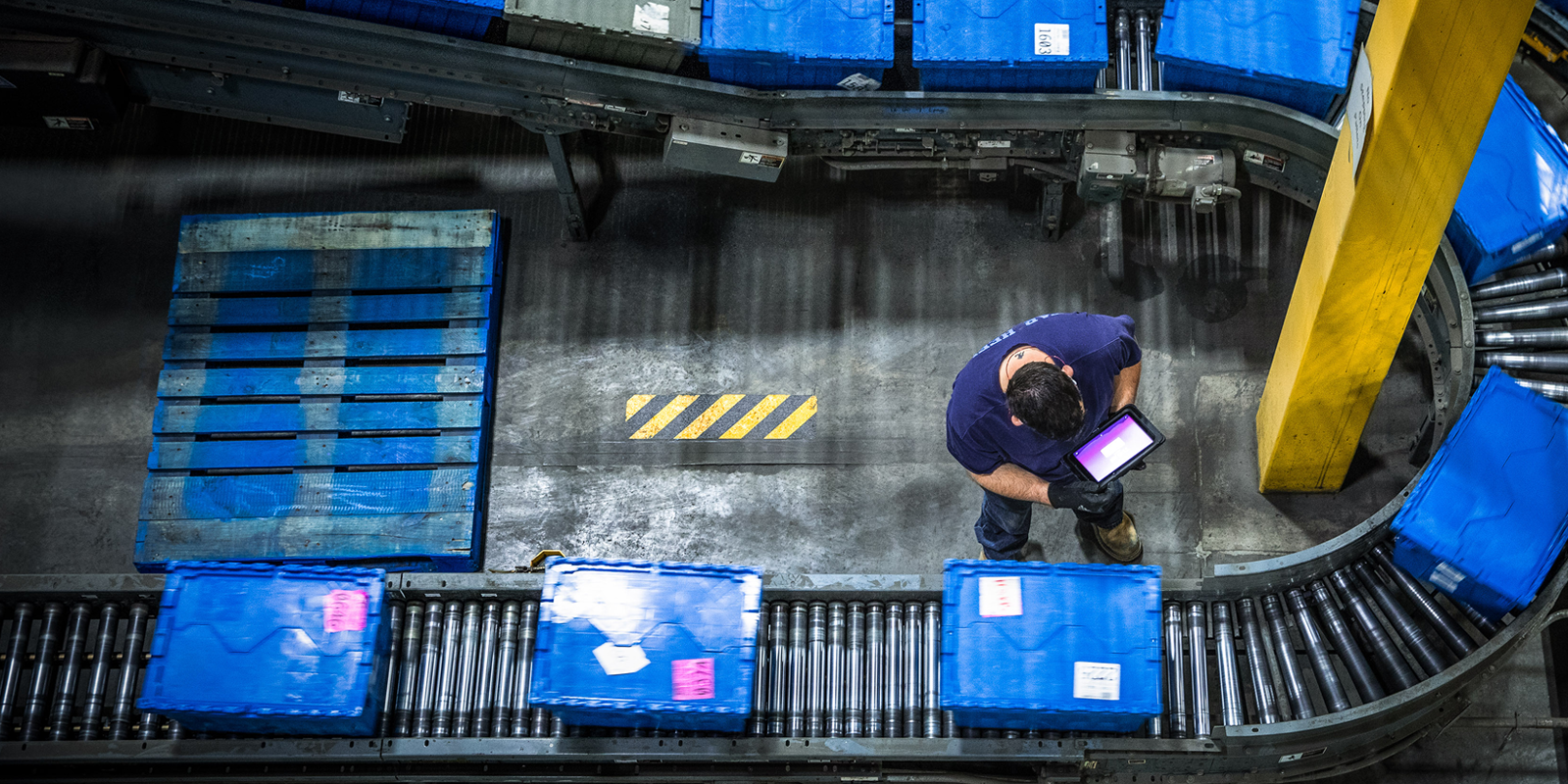 Bird’s-eye-view of a man working in a warehouse while logging information into a tablet as packages travel on conveyor belts