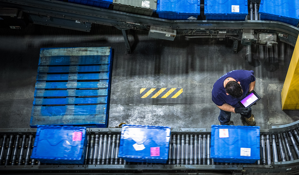 Bird’s-eye-view of a man working in a warehouse while logging information into a tablet as packages travel on conveyor belts