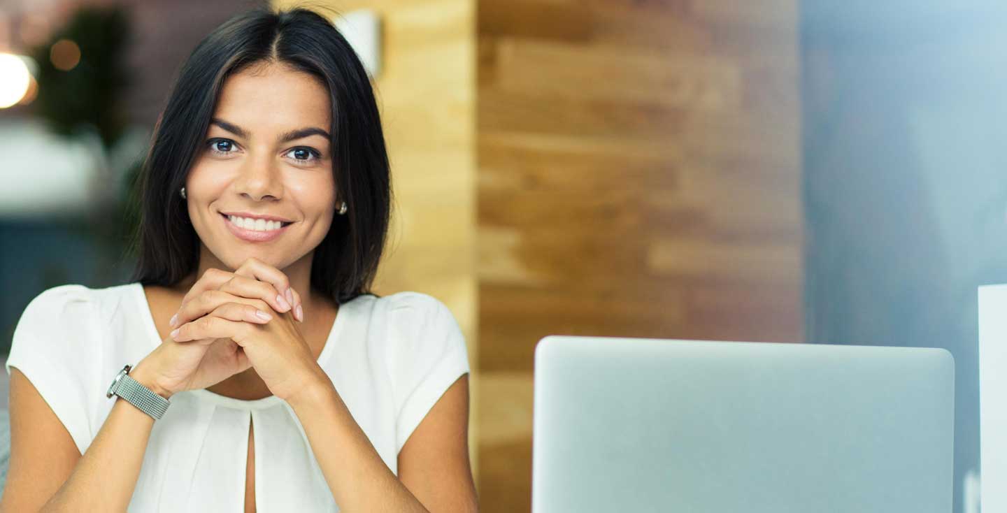 Woman sitting near a laptop looking at camera, folding her hands under her chin.