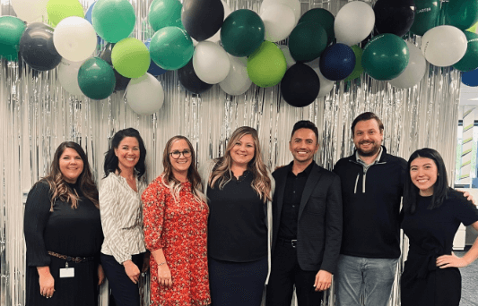Men and women Aston Carter employees posing under a balloon wall at an event.