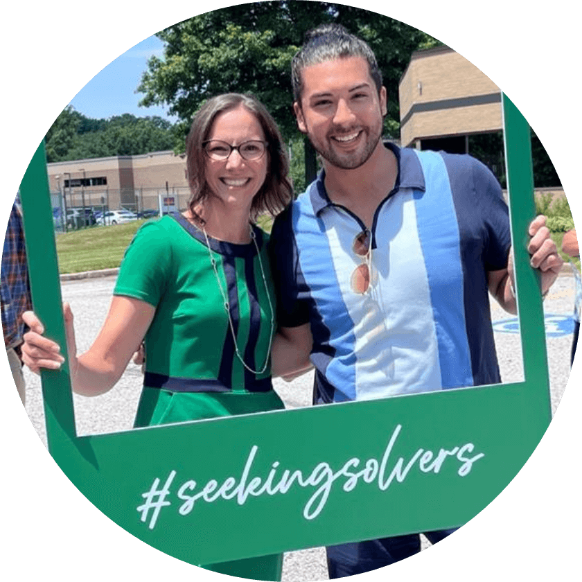 A man and woman Aston Carter employee posing behind a "seeking solvers" sign.