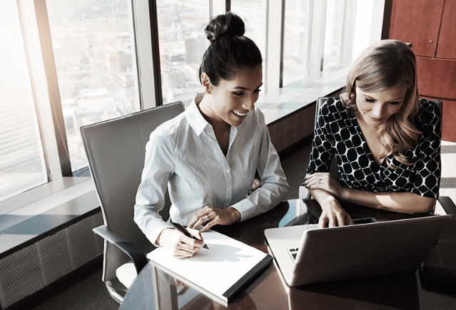 Two professional women sitting at a desk, talking, and working on a laptop.