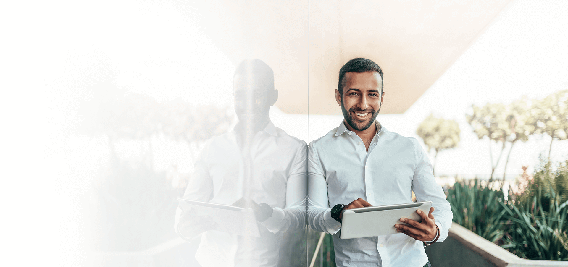 Professional leaning against a glass wall smiling and holding a tablet device.