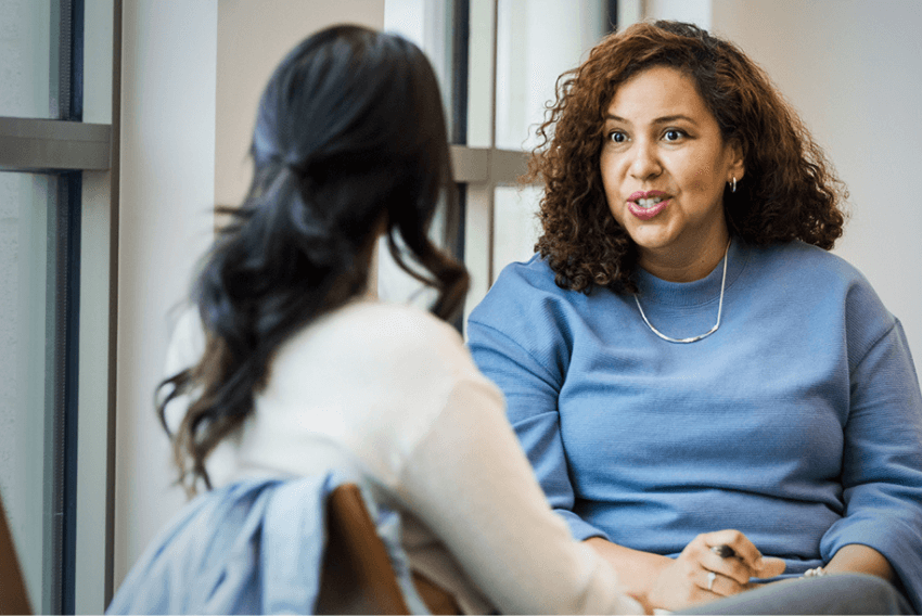 A woman with curly hair in a blue sweater talks to another woman seated across from her.