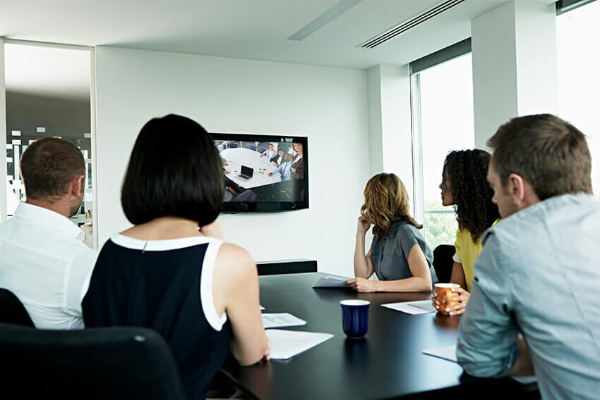 Five diverse empolyees sitting at a conference table on a video call looking at the TV screen 