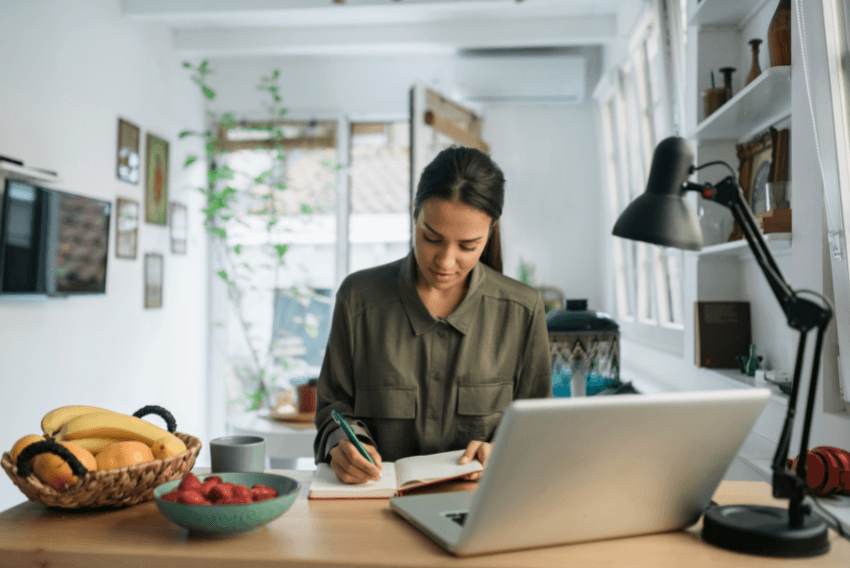 Female employee taking notes while in a virtual meeting from her kitchen table.