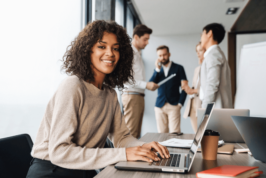 young woman working on laptop smiling at camera with colleagues in the background