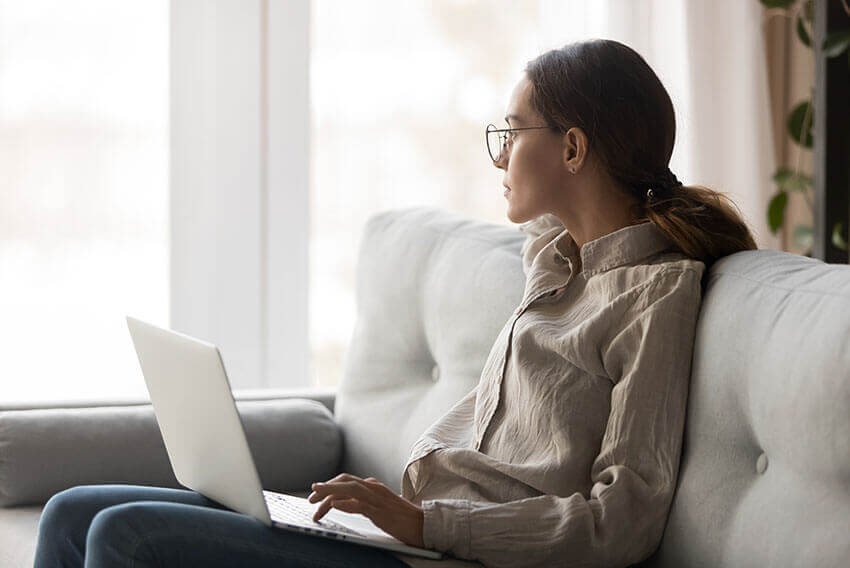 Woman sitting on couch with laptop in her lap, as she gazes out the window