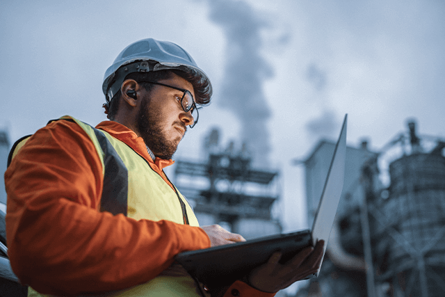 Man in hard hat and safety vest working on laptop outside energy facility