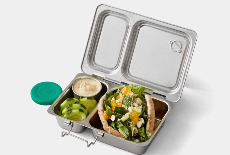 A silver steel bento box with two compartments, one compartment containing a cucumbers and a dip and the other containing a sandwich.